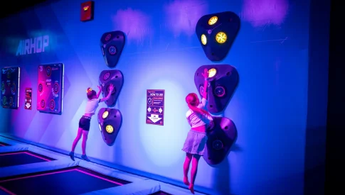 Twee jonge meiden springen op trampolines en raken oplichtende doelen op een verticale wand. Deze interactieve speeltoestellen in een trampolinepark dagen kinderen uit tot beweging onder sfeervolle paarse en blauwe neonverlichting.